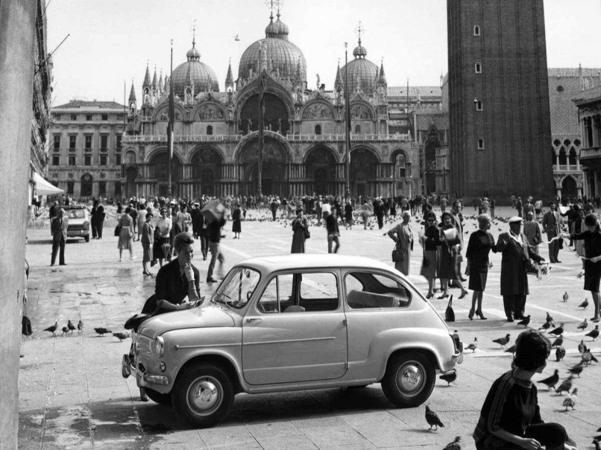 Fiat 600, de lado, estacionado na Praça de São Marcos, em Veneza, na Itália, com mulher debruçada sobre o capô; ao fundo, são vistas várias pessoas caminhando e a catedral, em foto preto e branca