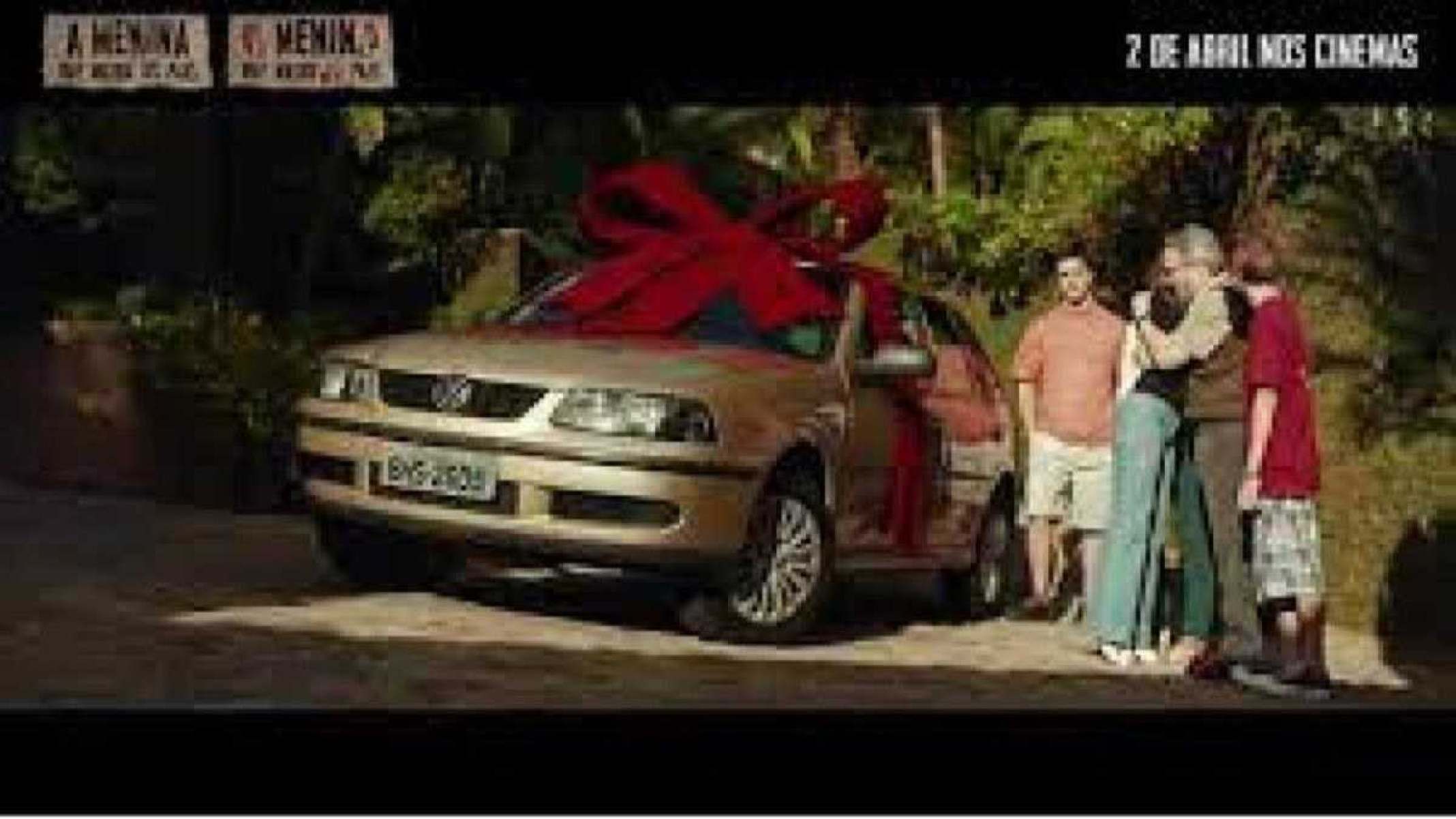  Cena do filme sobre o crime "a garota que matou os pais" Volkswagen gol amarelo envolto por um laço vermelho estacionado com jardim no fundo. Na lateral uma mulher loira de calça jeans azul abraçando um homem de roupa marrom enquanto dois rapazes aparecem ao fundo observando 