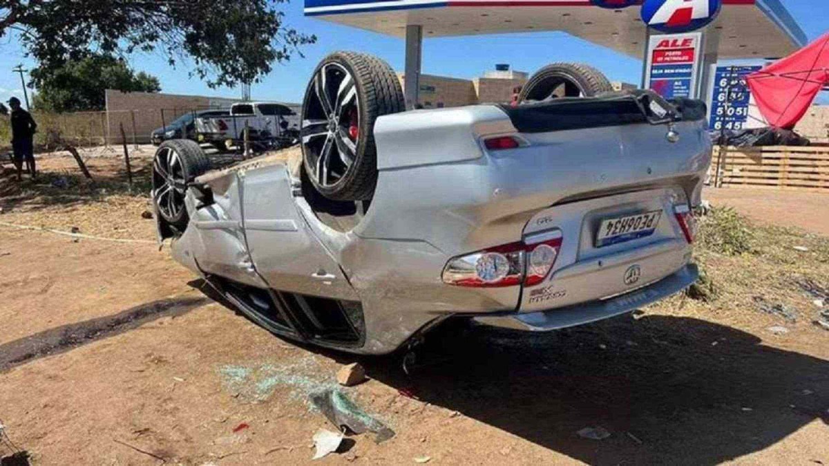 Fotografia de veiculo na cor cinza com as 4 rodas para cima e sinais de destruição em frente a um posto de gasolina