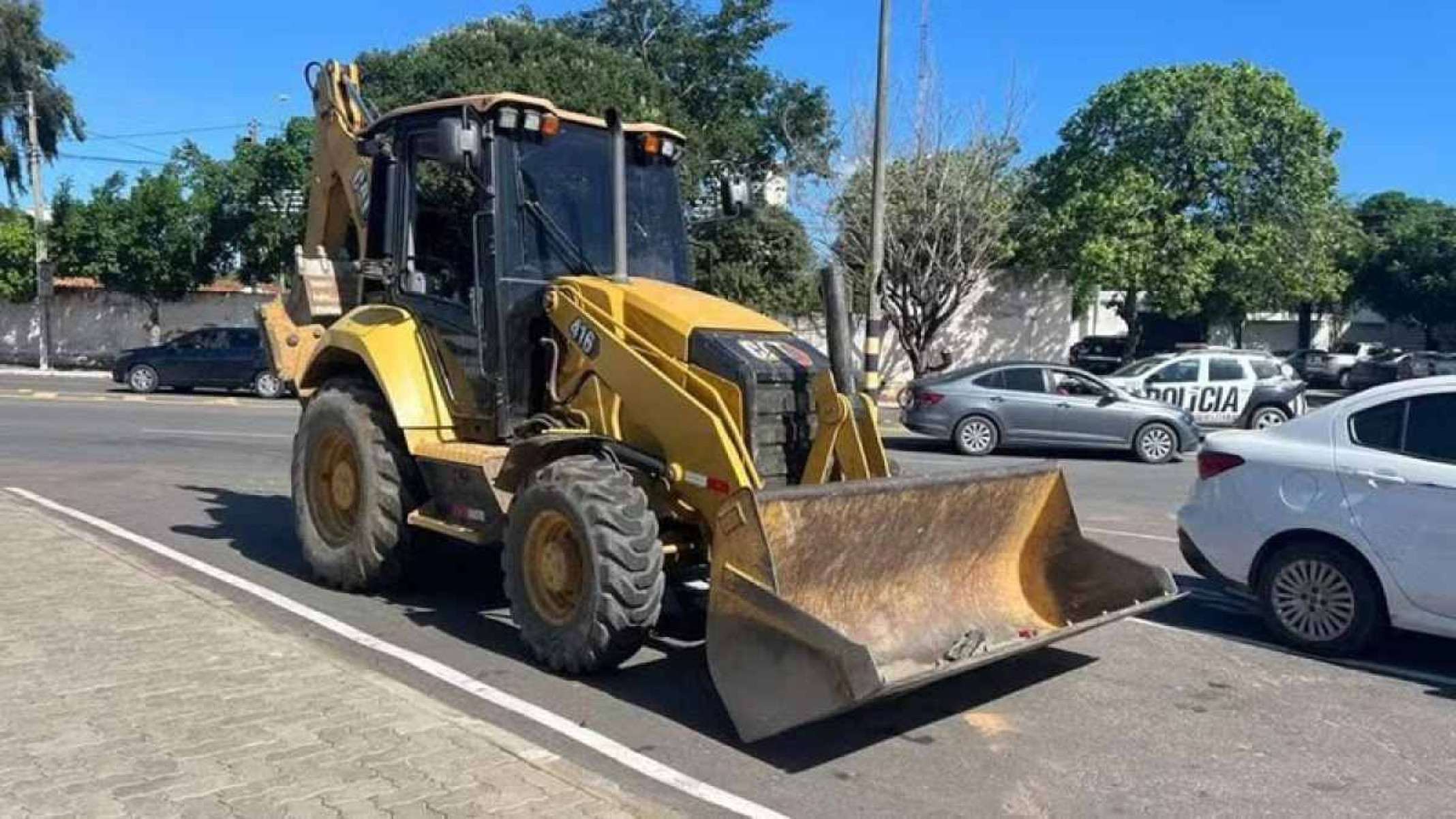 Trator amarelo estacionado em uma rua onde carros passam ao fundo