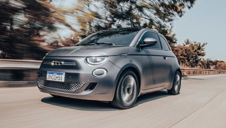 Fiat 500e modelo 2021 hatch el&eacute;trico cinza de frente em movimento no asfalto com mato ao fundo