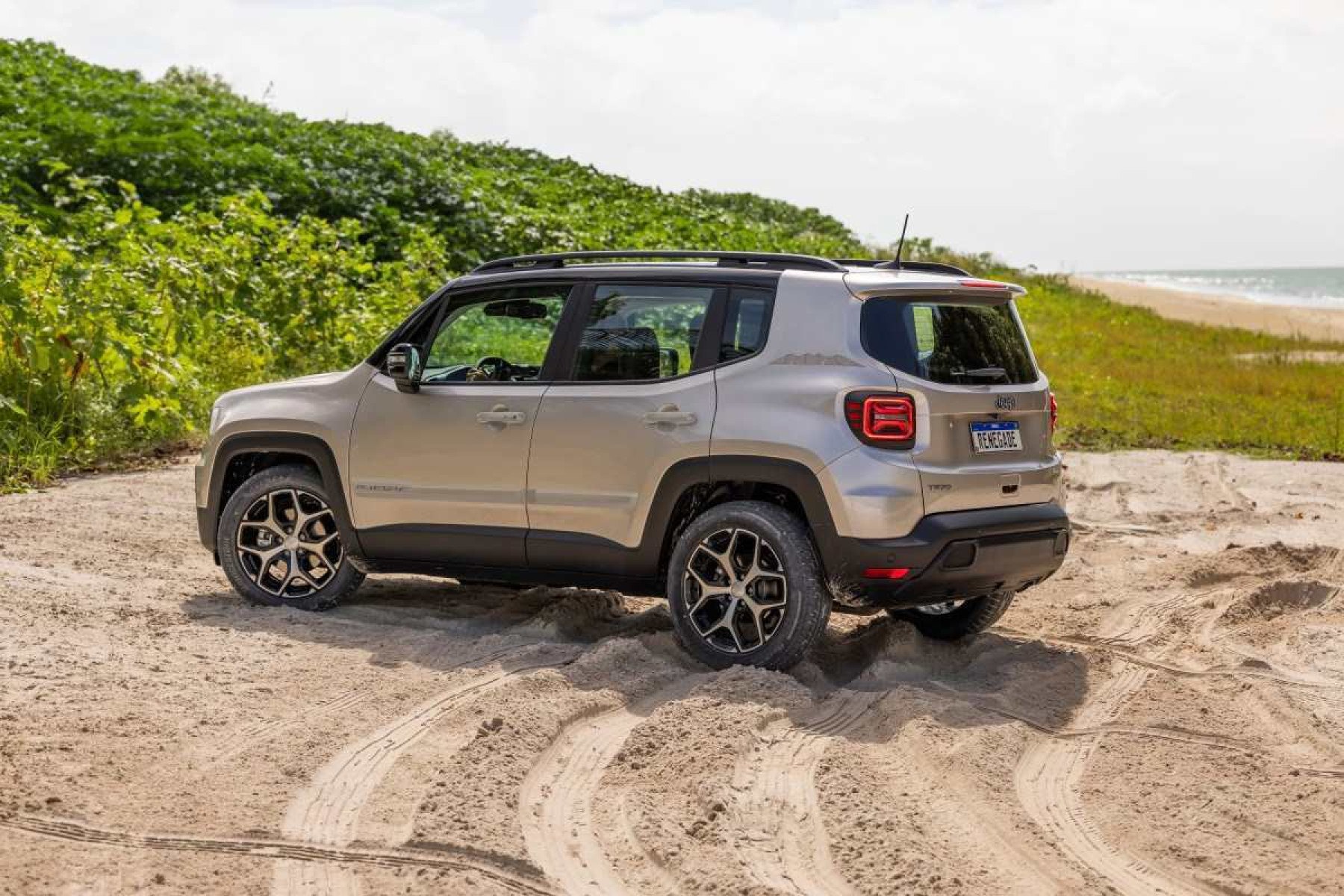 Jeep Renegade Sahara modelo 2025 cinza com teto preto de lateral traseira na areia com mato ao fundo