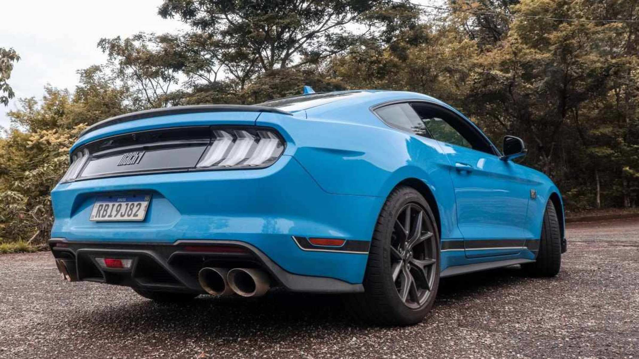 Ford Mustang azul de traseira estático no asfalto com mata ao fundo