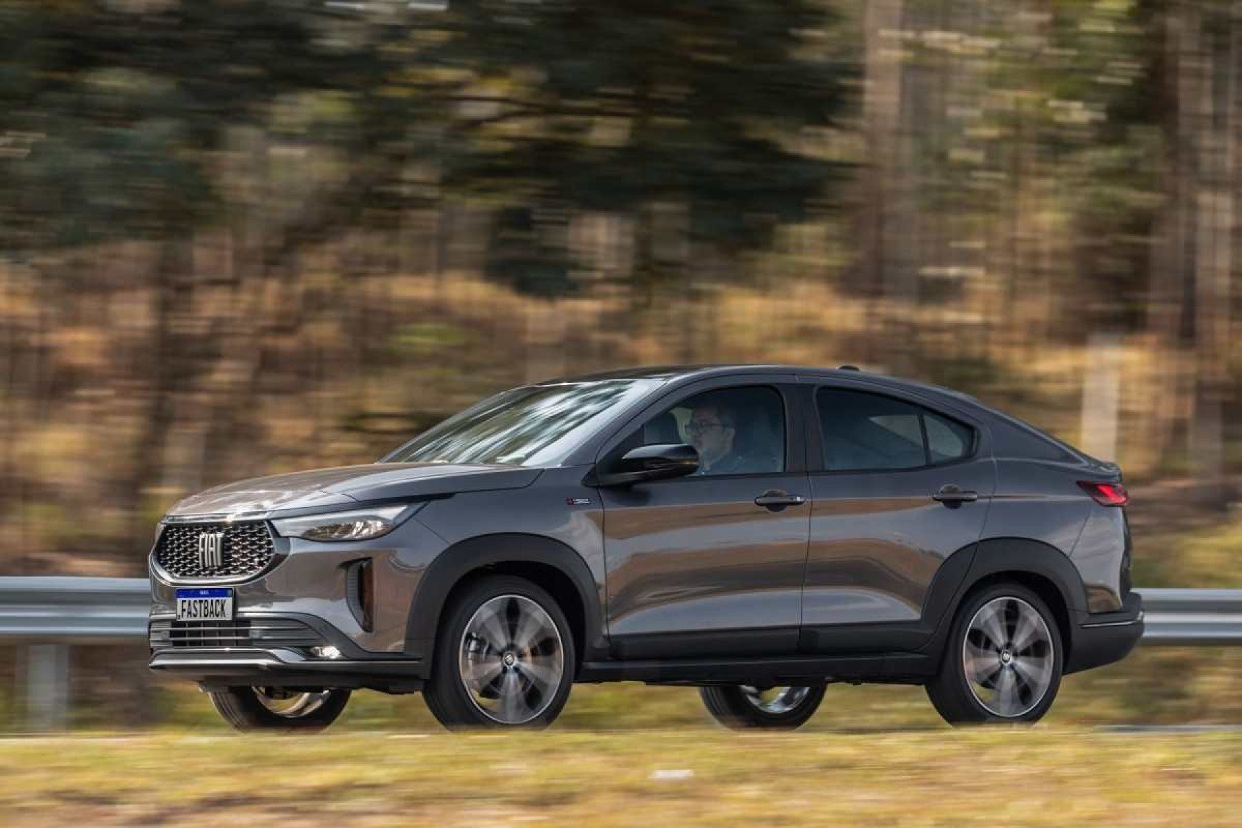 Fiat Fastback modelo 2023 cinza escuro de lateral em movimento no asfalto com mato ao fundo