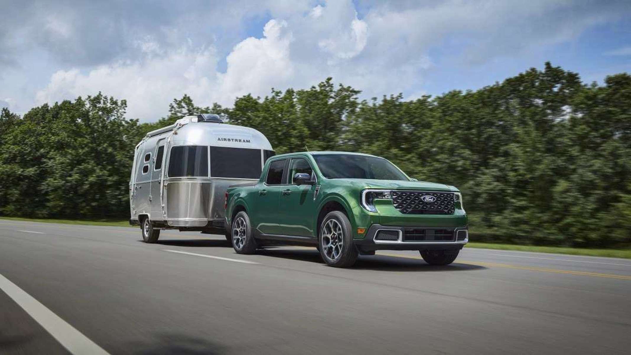 Ford Maverick 2025 verde de frente puxando um pequeno trailer é visto de frente com detalhes no lado direito.  Veículo é visto viajando em uma rodovia pavimentada com árvores e folhagens escuras e um céu claro cheio de nuvens ao fundo