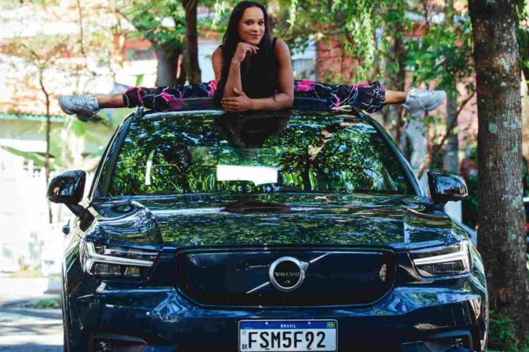 Rebeca Andrade abrindo o teto de um Volvo EX40 preto visto de frente.  Ao fundo você pode ver uma rua pavimentada com árvores na calçada, a imagem está desfocada