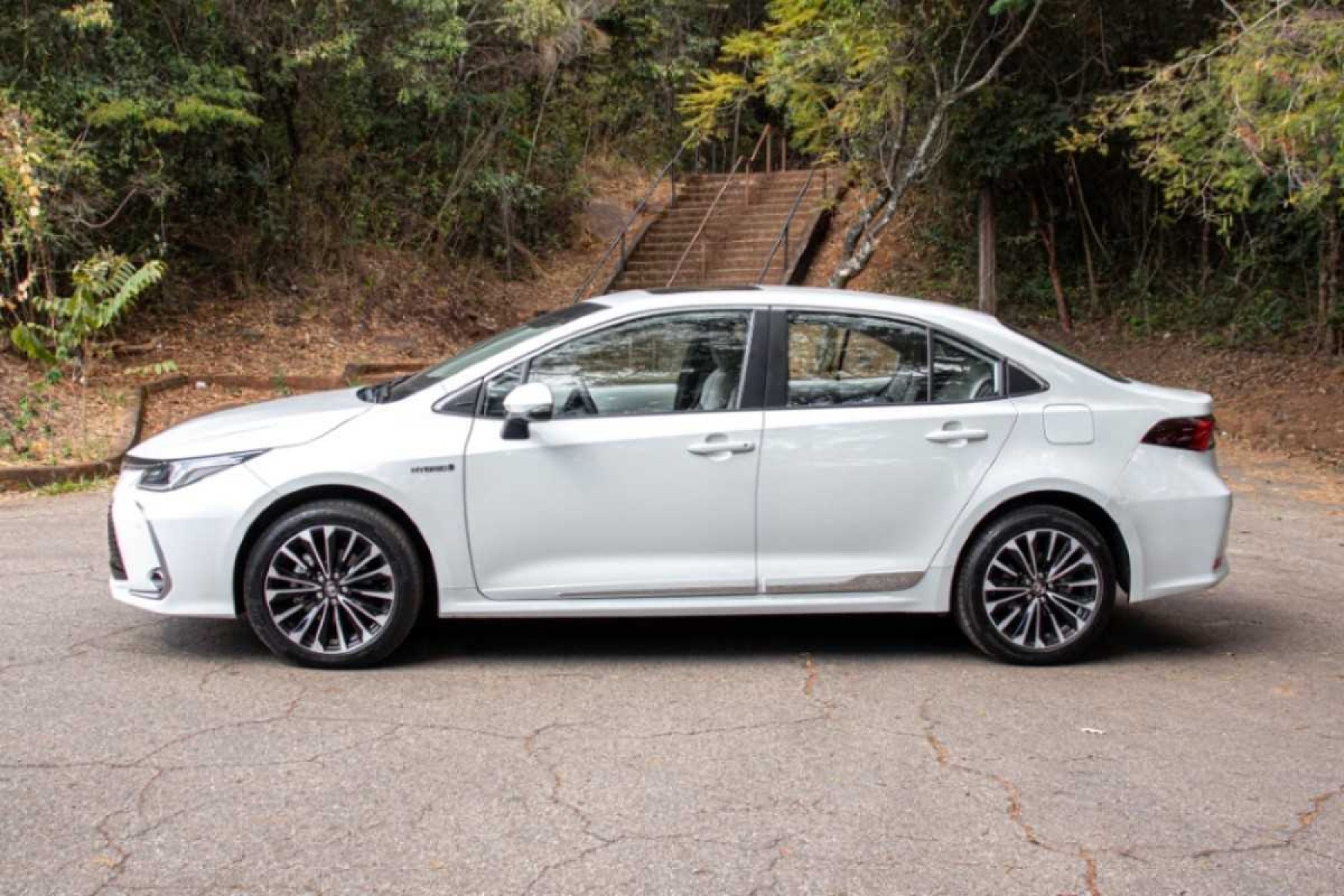 Toyota Corolla Altis Hybrid Premium modelo 2024 branco de lateral no asfalto com escadaria e mata ao fundo