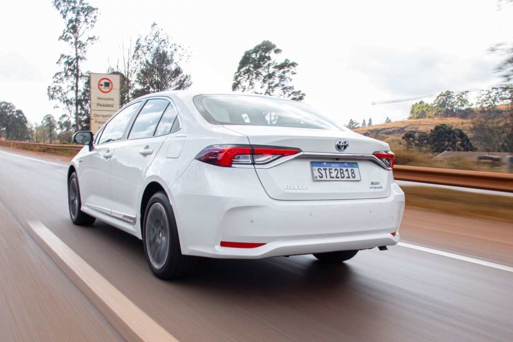 Toyota Corolla Altis Hybrid Premium modelo 2024 branco de traseira em movimento no asfalto com mata ao fundo