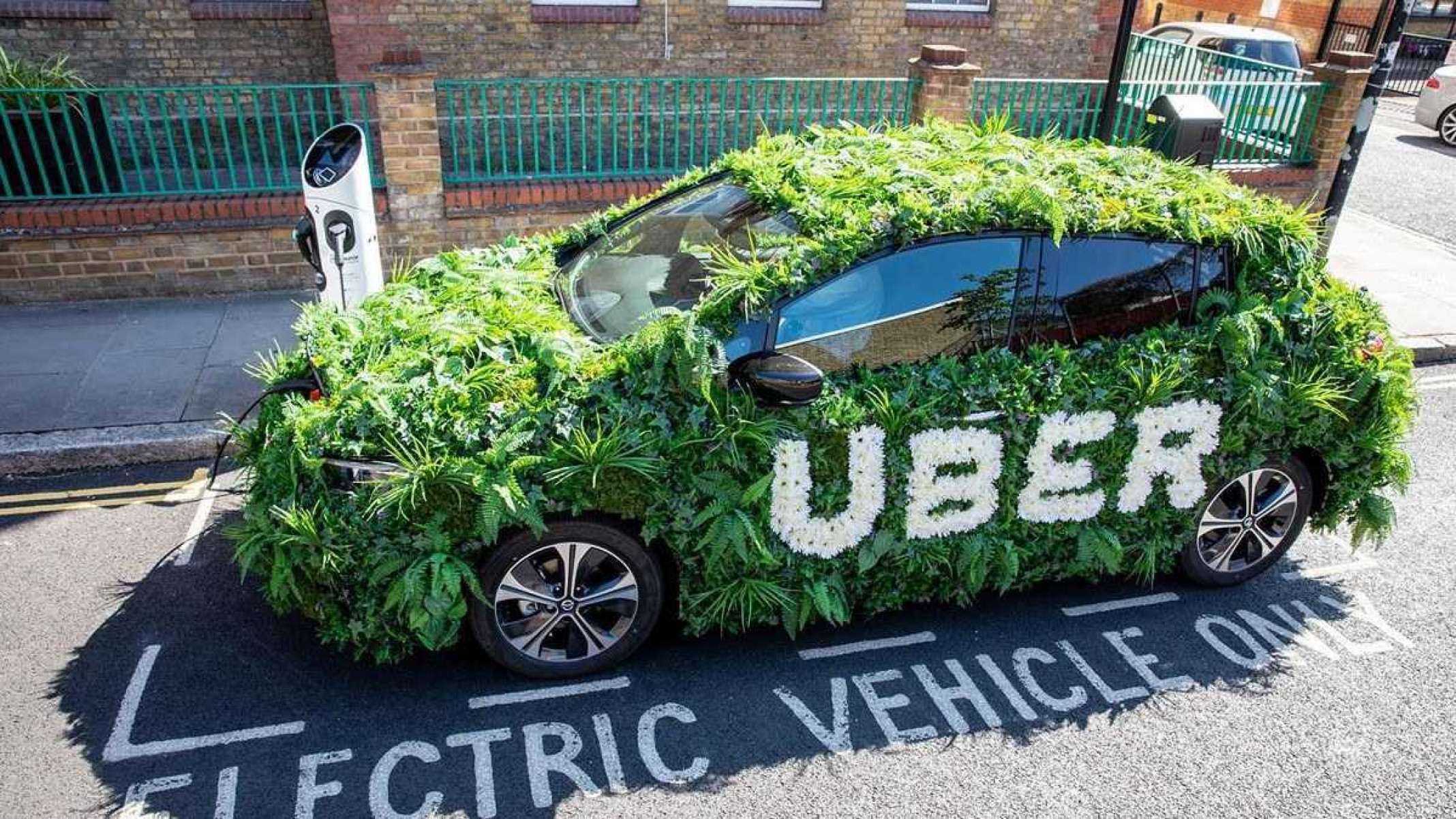 Carro elétrico coberto de planta com o nome "Uber" na lateral estacionado em rua asfaltado com prédio ao lado