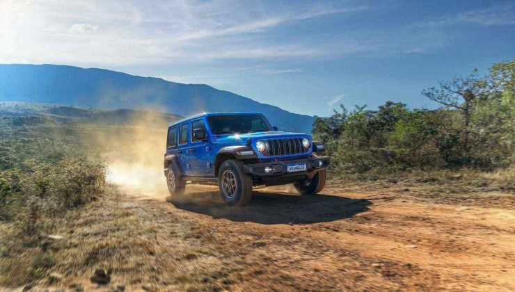 Jeep Wrangler Rubicon 2025 azul visto pela dianteira está trafegando em chão de terra vermelha e subindo poeira. Ao lado é possível ver vegetação rasteira com céu claro com nuvens ao fundo
