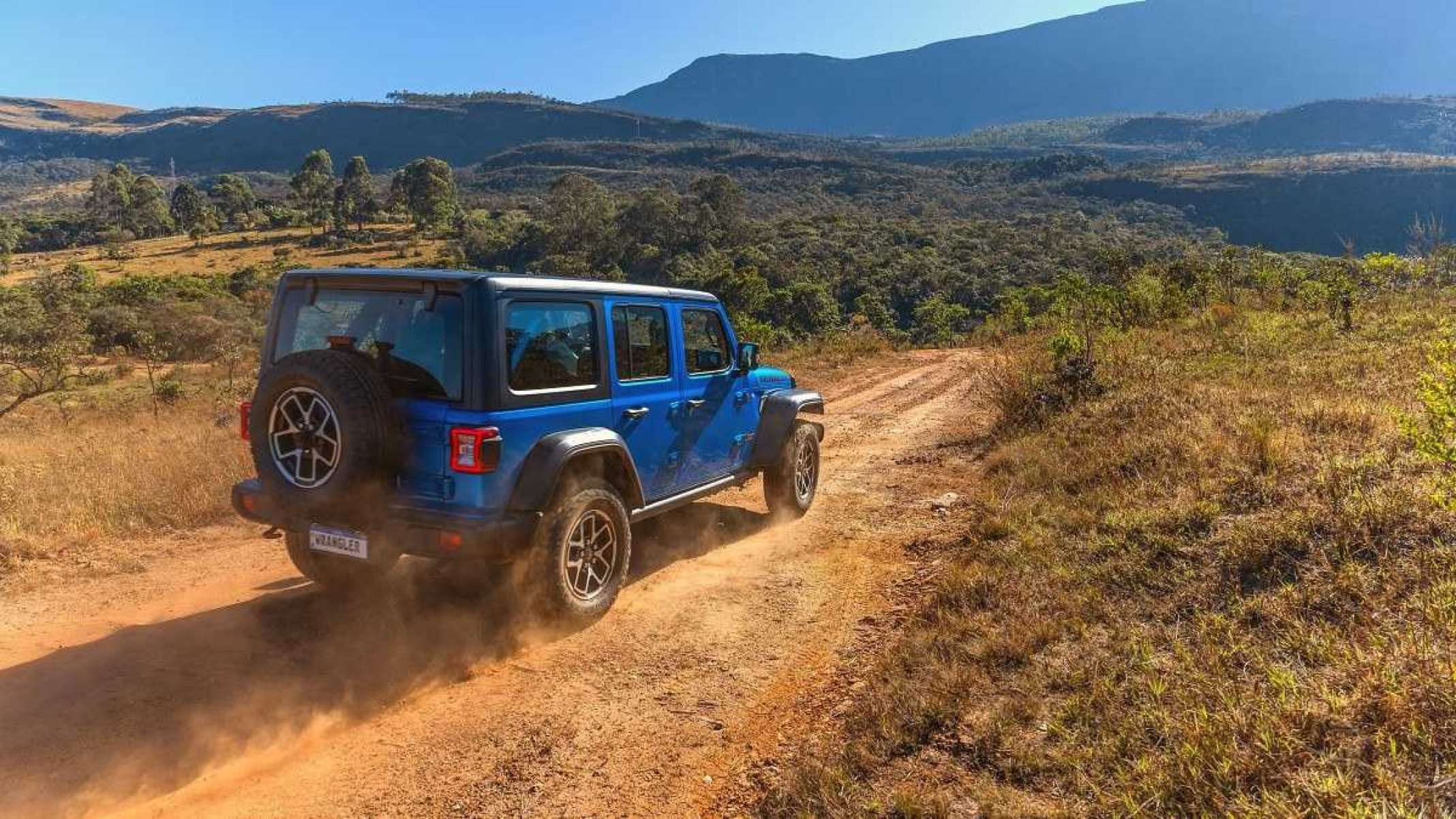 Jeep Gladiator Rubicon 2025 azul visto pela traseira com detalhes da lateral direita. Veículo trafega em chão de terra e levanta poeira. Ao fundo é possível ver montanha coberta por vegetação e céu claro.