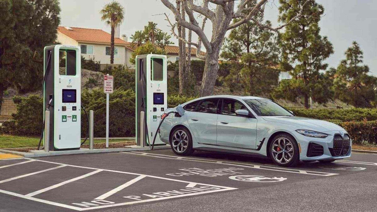 Carro elétrico branco parado em estação de carregamento com árvores ao fundo