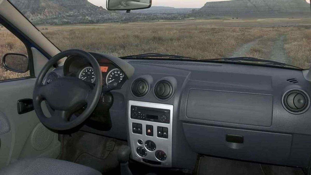 Interior de Renault Logan de primeira geração com acabamento em preto e detalhes em branco. Pelo para-brisas é possível ver gramado seco com céu nublado acima. 