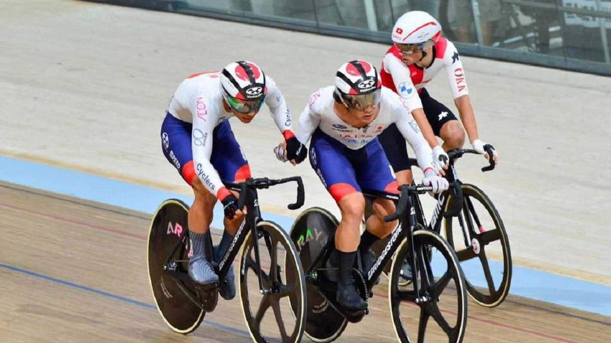 Três atletas japoneses com collants branco, azul e vermelho estão montados em V-Izu TCM2. Atletas estão andando com as bicicletas em pista utilizada nas Olimpíadas de Paris 2024