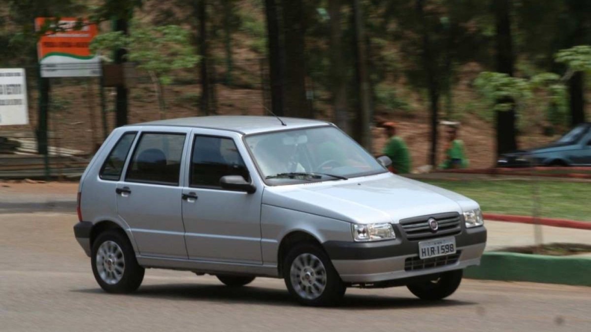 Fiat Uno Mille Fire Economy Flex modelo 2009 quatro portas prata de frente em movimento no asfalto com praça ao fundo