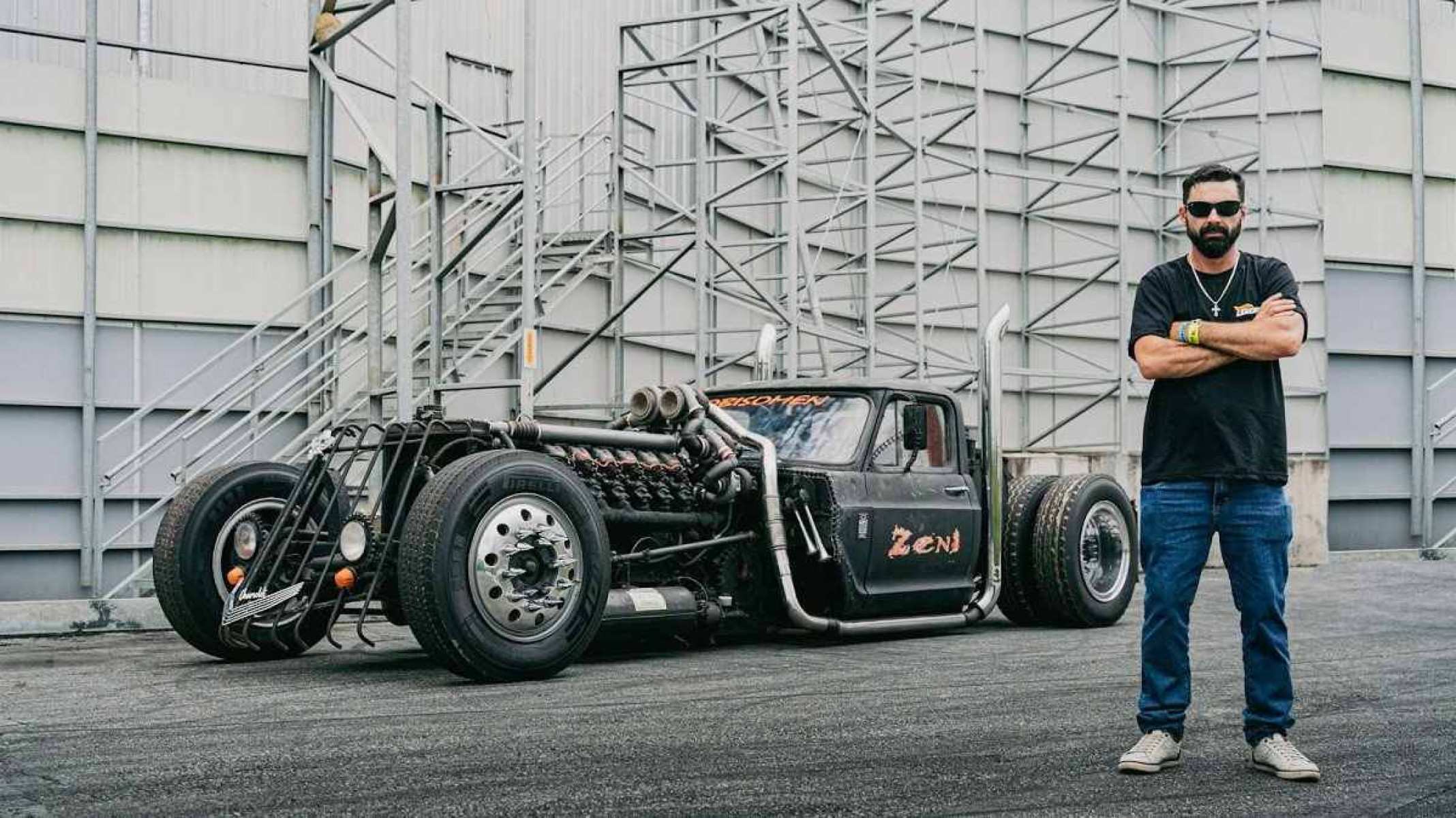 Pessoa com cabelo preto curto e longa barba escura veste uma camisa preta com jeans azul escuro. O homem está localizado no lado direito da fotografia e posa ao lado de um Chevrolet V12 transformado. Ao fundo há grandes silos cinzentos. 