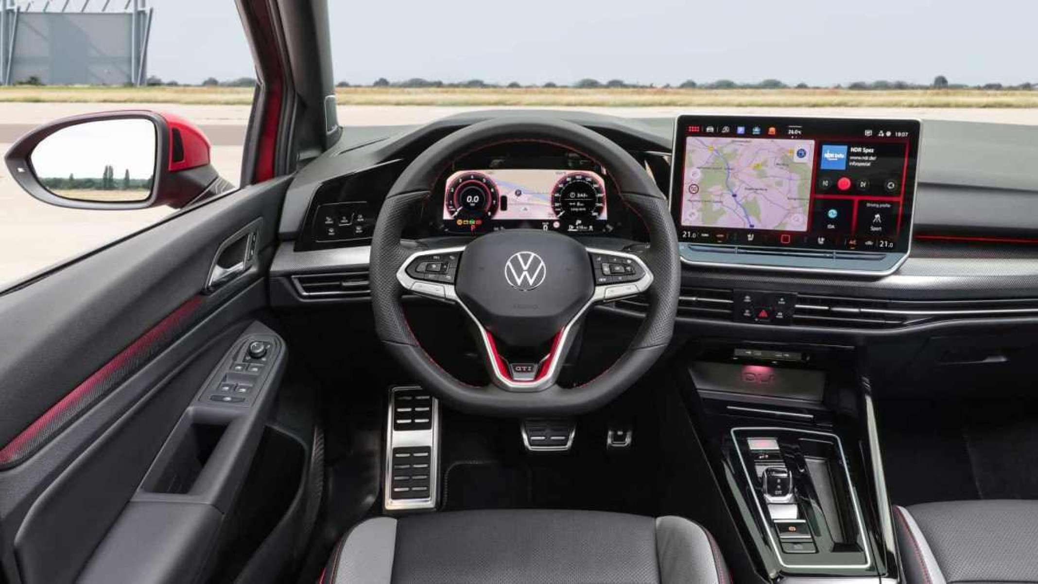 Interior do W Golf GTI com telas acesas e acabamento em preto com pequenos detalhes vermelhos no volante. Pelo para-brisa e pela janela do motorista é possível ver uma estrada asfaltada com grama ao fundo.