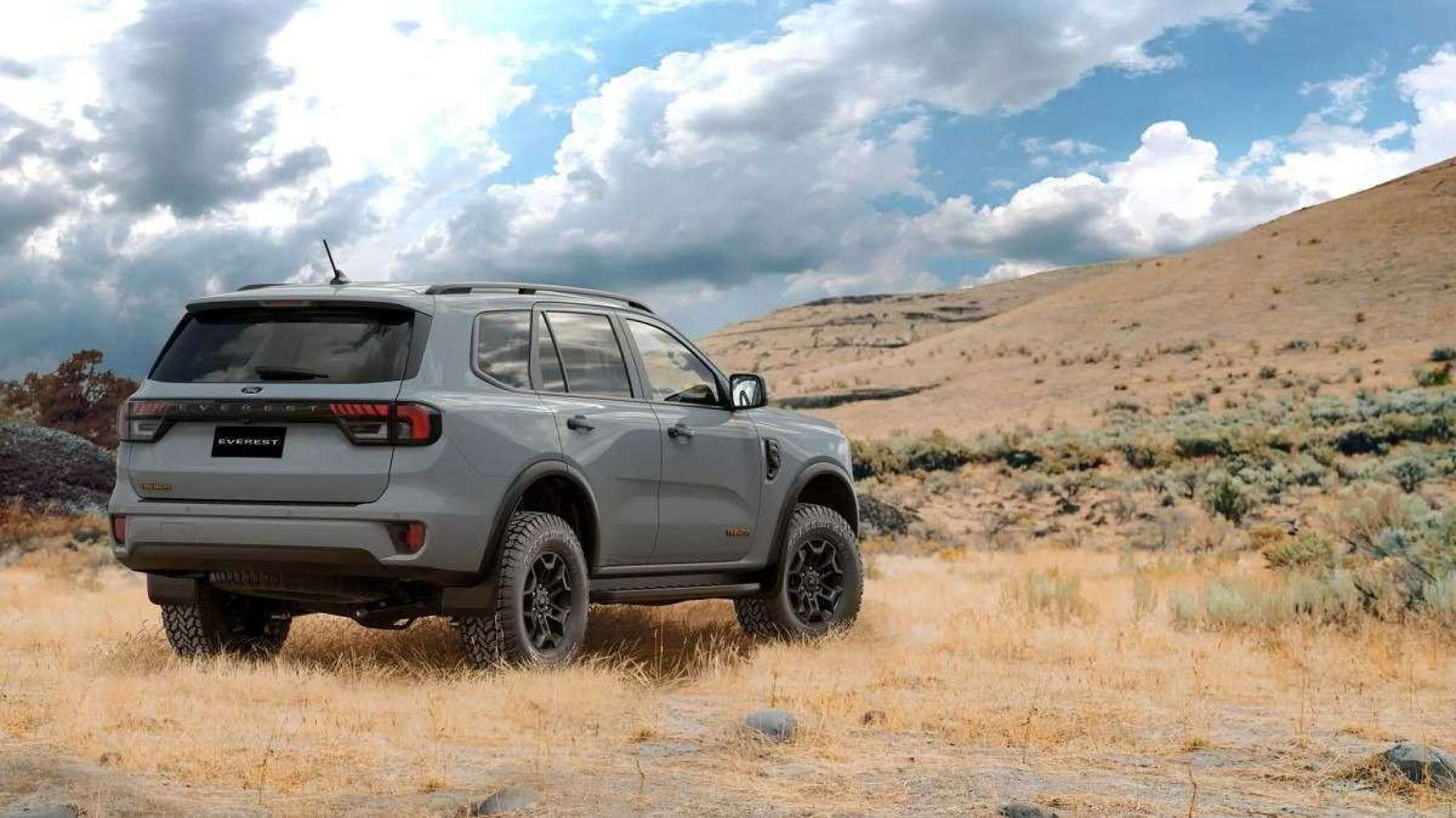 Ford Everest Tremor cinza visto pela traseira com detalhes da lateral direita. Modelo está estacionado em gramado de vegetação seca com montanha vista ao fundo e céu claro com nuvens