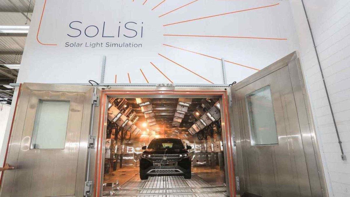 Porta do quarto na fábrica da Volkswagen. A parede é branca com o logo ‘SoLiSi’ em linhas pretas e amarelas, que simulam raios de Sol, na lateral. É possível ver seu interior, que contém o veículo da fabricante com diversas luzes laterais.