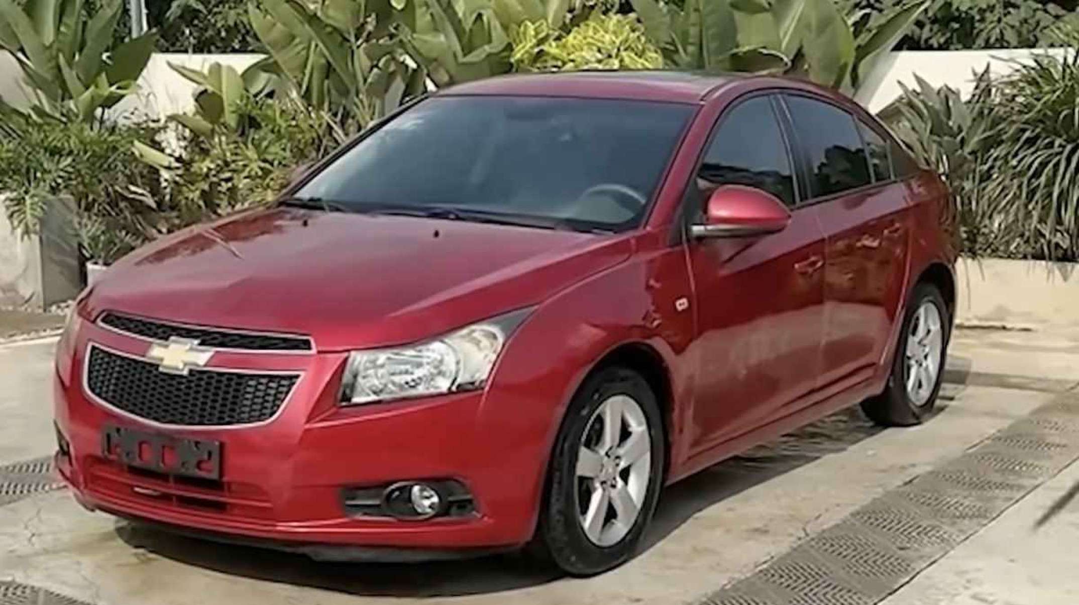 Chevrolet Cruze vinho é visto de frente com detalhes na lateral esquerda. O veículo está estacionado em terreno cinza. Ao fundo você pode ver algumas folhas verdes escuras.