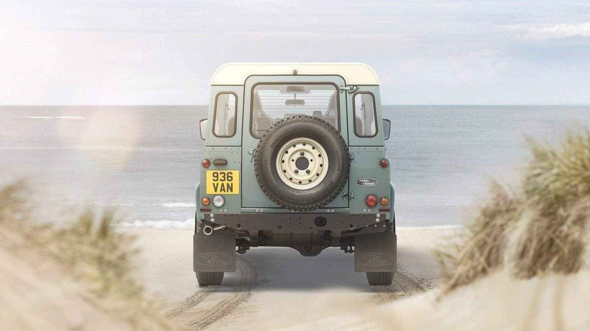 Traseira de Land Rover Defender na cor verde com teto branco. Veículo possui pneu na parte de trás. Veículo está parado em praia e mar e céu claro é isto ao fundo.