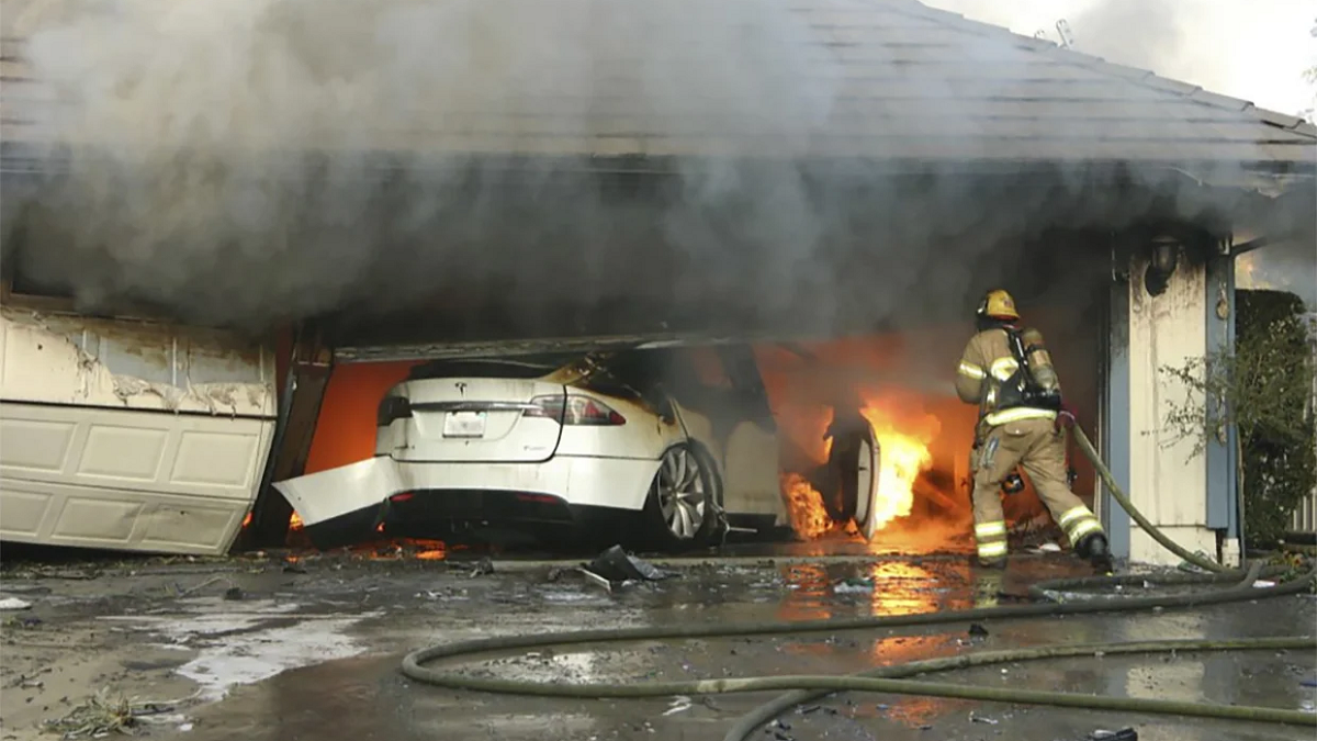 Carro Tesla cinza dentro da garagem em chamas com bombeiros tentando apagá-lo