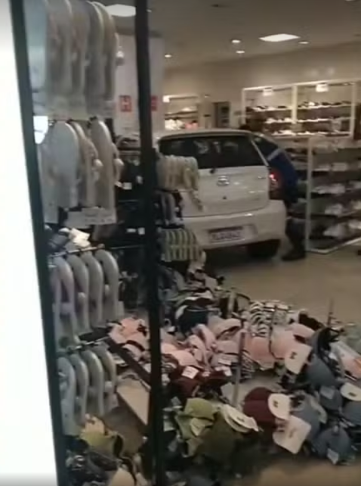 Toyota Etios branco dentro de uma loja de departamento batido em uma pilastra, rodeado por prateleiras e roupas jogadas ao chão e um homem tentando resgatar uma criança que está na parte de trás do carro